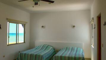 A room with two beds, a ceiling fan, a window with a view of the ocean, and a door.