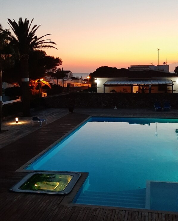 A swimming pool at dusk with a sunset in the background, a palm tree, and a building with lights on. - BEAUTIFUL DUPLEX IN CALA SANTANDRIA-CIUTADELLA WITH POOL AND WIFI (Cala Santandria-Ciutadella-Islas Baleare)