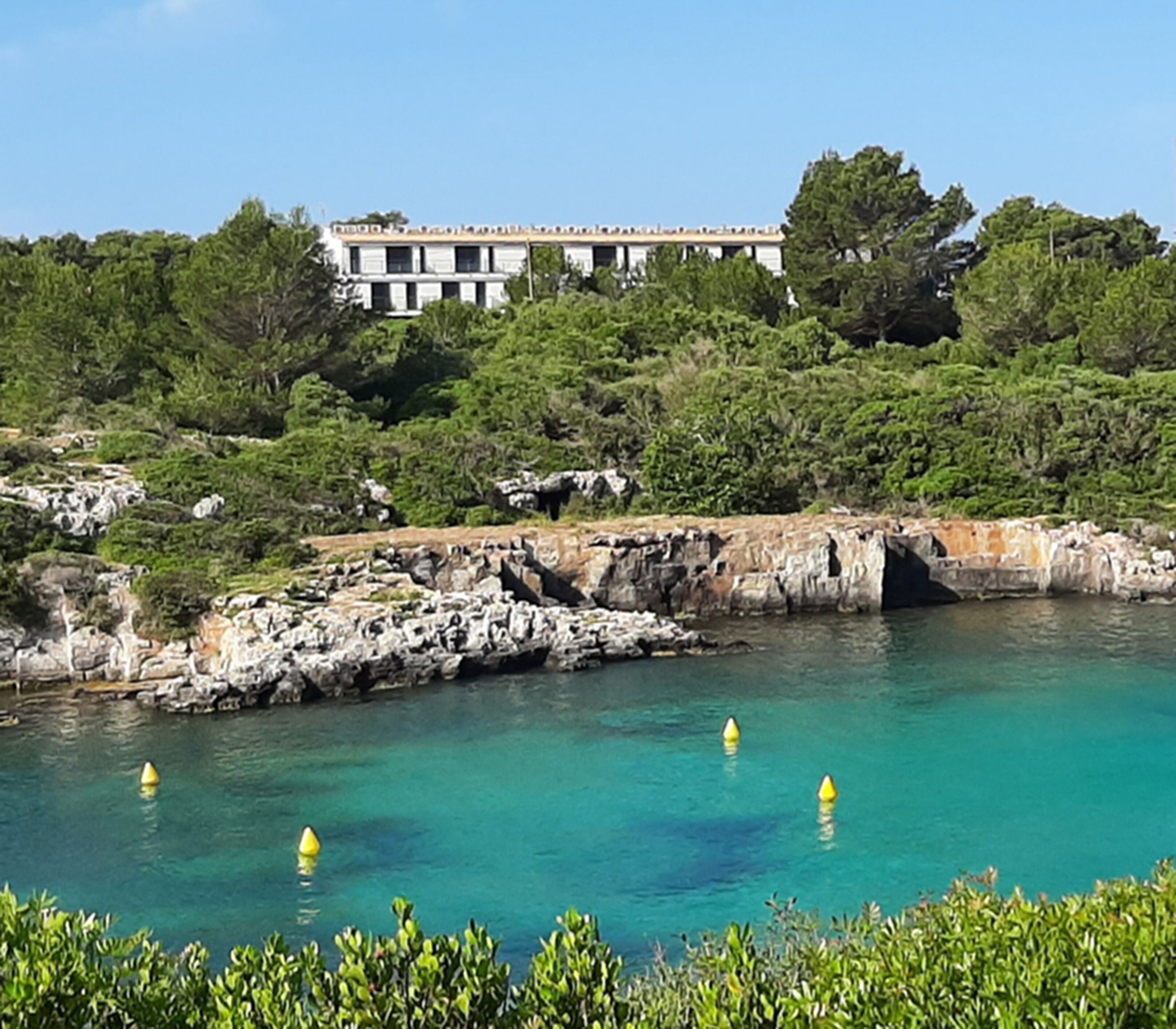 A coastal hotel with a rocky shoreline, clear turquoise water, and lush greenery.