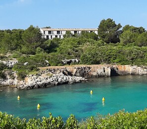 A coastal hotel with a rocky shoreline, clear turquoise water, and lush greenery. - BEAUTIFUL DUPLEX IN CALA SANTANDRIA-CIUTADELLA WITH POOL AND WIFI (Cala Santandria-Ciutadella-Islas Baleare)