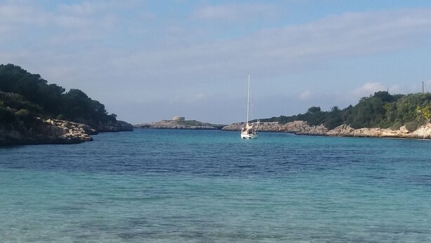 A beach with clear turquoise water, a sailboat in the distance, and rocky hills in the background. - BEAUTIFUL DUPLEX IN CALA SANTANDRIA-CIUTADELLA WITH POOL AND WIFI (Cala Santandria-Ciutadella-Islas Baleare)
