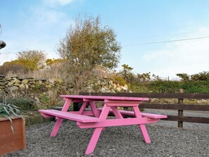 Una mesa de picnic rosa en un jardín, con un muro de piedra y una valla de madera al fondo.