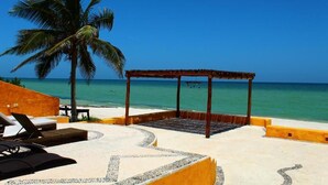 Un área de descanso junto a la playa con un pabellón, sillas de salón y una palmera.