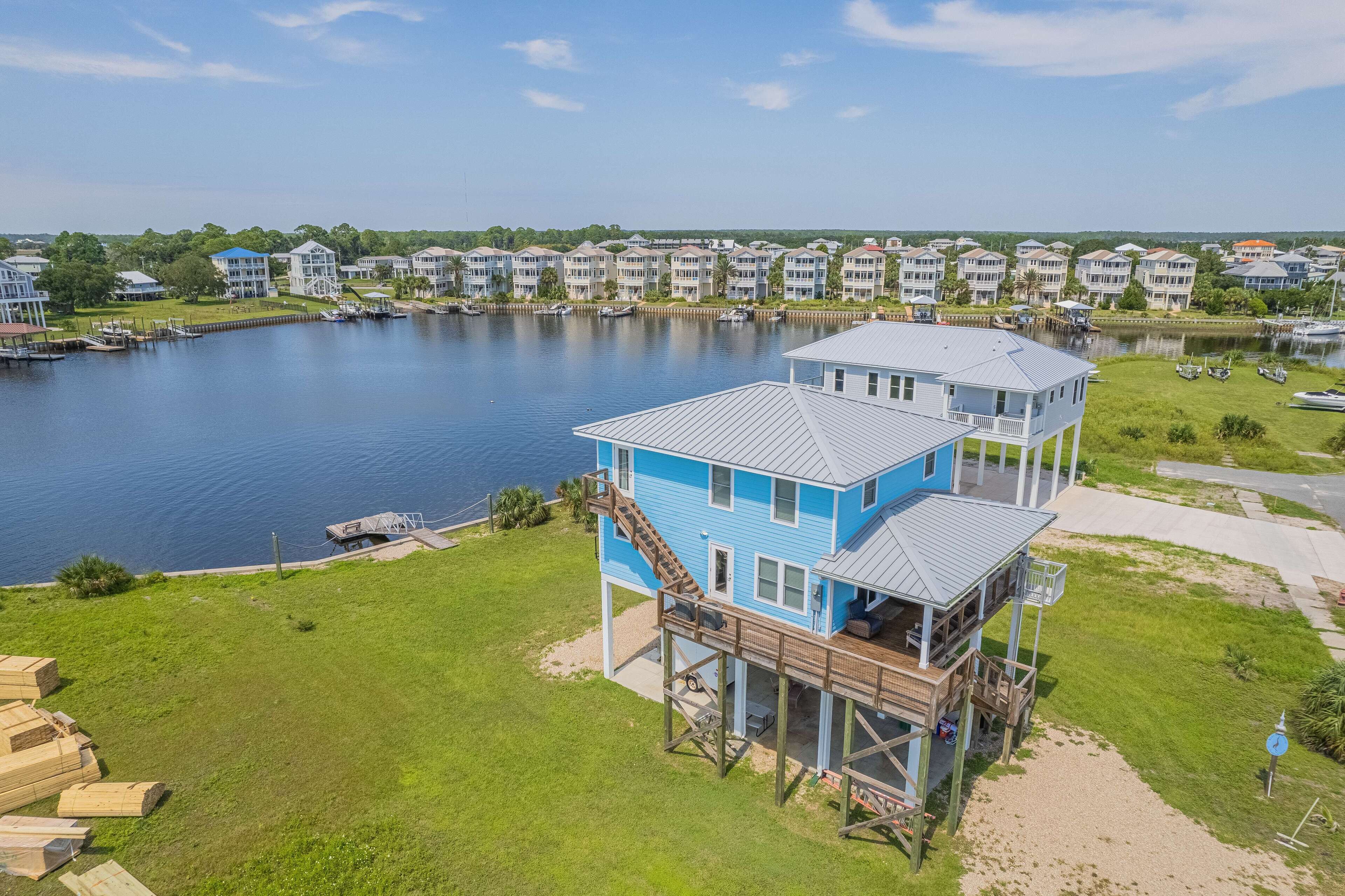 A two-story house with a deck and a staircase leading to the second floor, situated by a lake with other houses in the background.