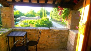 Un patio con paredes de piedra, una mesa y dos sillas, con vistas a un jardín con un sendero y árboles.