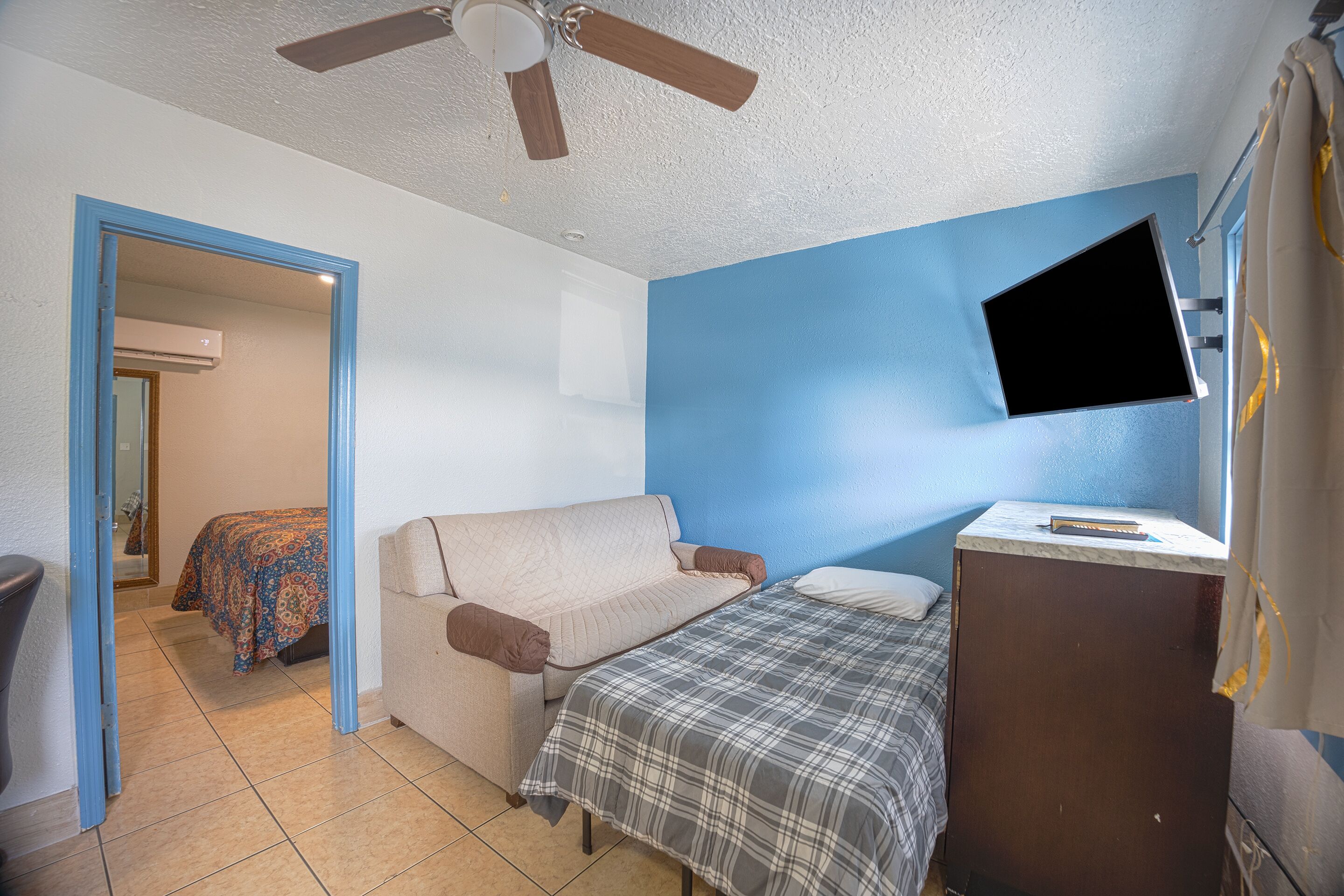 A room with a bed, a desk, a ceiling fan, and a TV mounted on the wall.