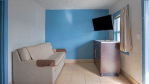 A room with a blue wall, a beige sofa, a TV mounted on the wall, and a window with curtains. - Aladdin Motel By OYO Merritt Island (Merritt Island)
