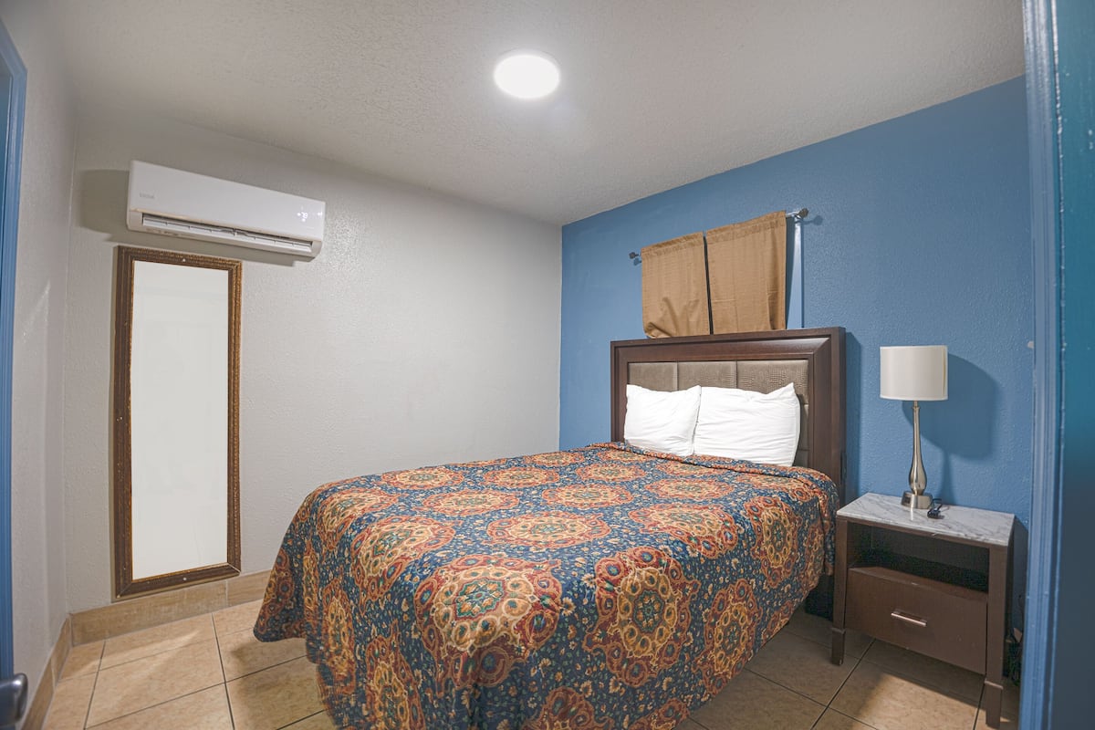 A hotel room with a bed, a nightstand, a lamp, an air conditioner, a window with curtains, and a door.