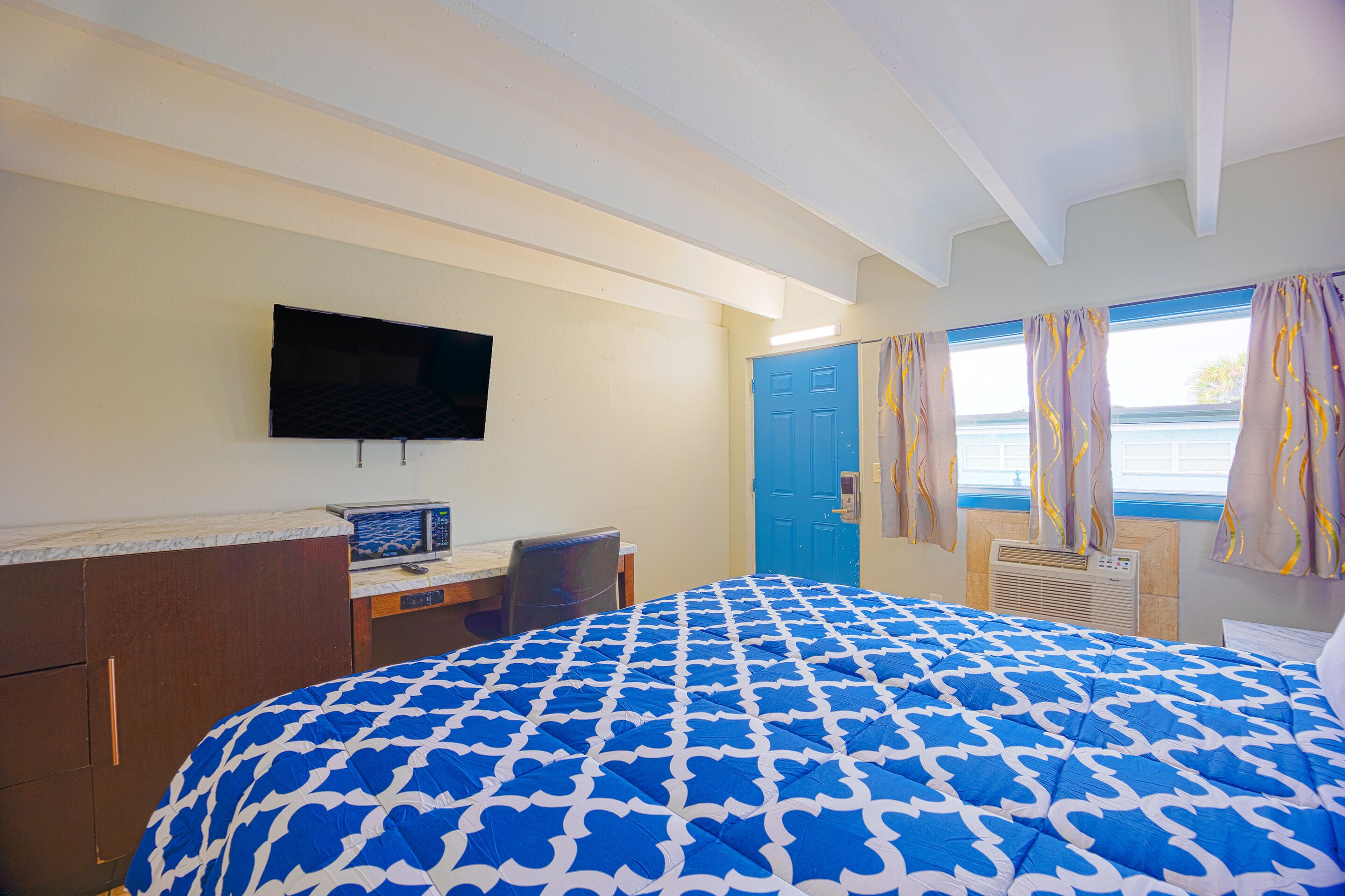 Habitación de hotel con cama, escritorio, silla, televisor, puerta azul y ventana con cortinas.