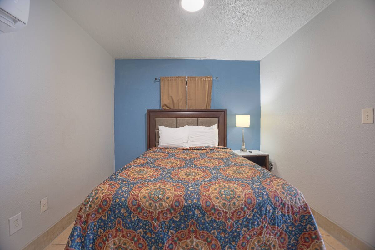 A bedroom with a patterned bedspread, a wooden headboard, a nightstand with a lamp, and a window with curtains.