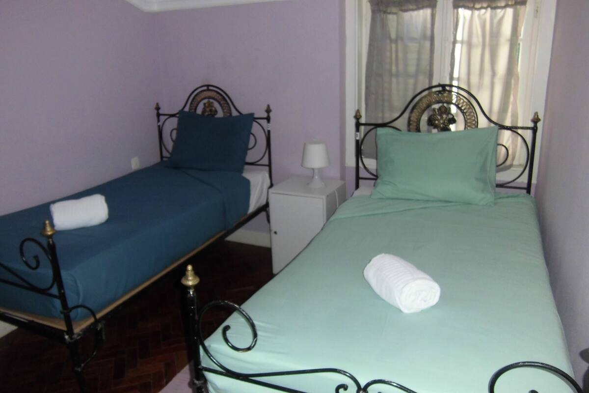 Two single beds with metal frames, a white nightstand, and a window with curtains.