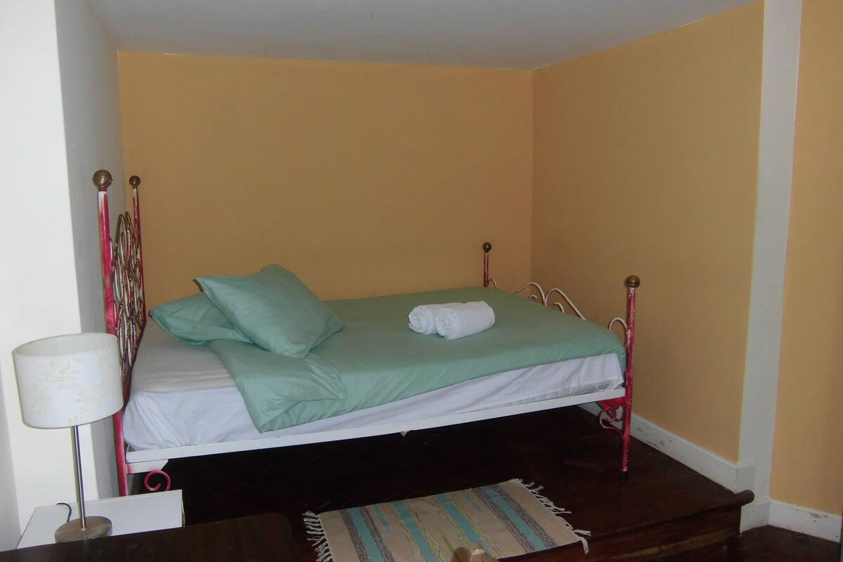 A single bed with a green bedspread and white linens, a small bedside table with a lamp, and a striped rug on the floor.