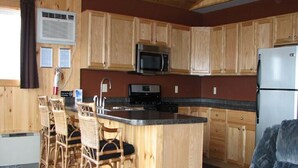 A kitchen with wooden cabinets, a microwave, a stove, and a refrigerator. There are wicker chairs around a bar area. - Cottage #13 - 3 bedroom/ 1 bath (Cape Vincent)
