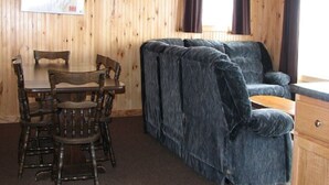 A wooden cabin interior with a dining table, chairs, a sofa, and a window with curtains. - Cottage #13 - 3 bedroom/ 1 bath (Cape Vincent)