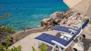 A beachside view with clear turquoise water, rocky terrain, and two sun loungers with a beach umbrella.