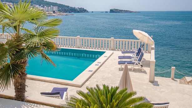 A poolside area with a view of the sea, lounge chairs, and a palm tree.