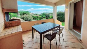 A balcony with a table and chairs, a grill, and a view of greenery and hills.