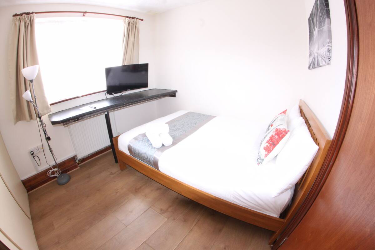 Ein Schlafzimmer mit einem Bett, einem Schreibtisch mit Computer, einem Fenster mit Vorhängen und einer Heizung.