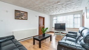 A living room with a black leather sofa, a wooden coffee table with a plant, a television, and a large window with blinds. - College Street Abode - Guest House (Portsmouth)