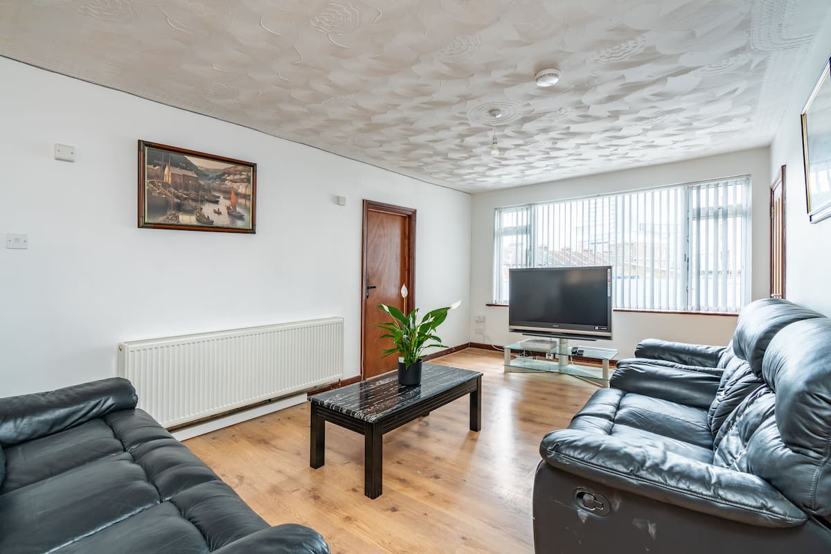 Ein Wohnzimmer mit einer schwarzen Ledersofa, einem Couchtisch mit Pflanze, einem Fernseher und einem großen Fenster mit Jalousien.