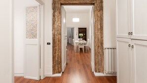 Un couloir doté d’un plancher en bois, de murs blancs et d’une grande porte encadrée en bois qui mène à une salle à manger avec une table et des chaises blanches.
