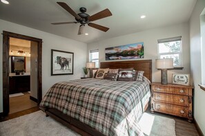 A bedroom with a large bed, a ceiling fan, a painting, and a nightstand with a lamp.
