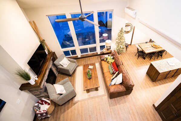 A spacious living room with a large window, a ceiling fan, a fireplace, a dining area, and a cozy seating arrangement.