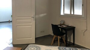 Una habitación con una mesa de madera, una silla negra, un tapete estampado, una puerta blanca y una ventana con persianas.