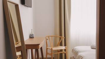 Un dormitorio con una cama, una silla de madera, una mesita con un florero y un televisor en la pared.