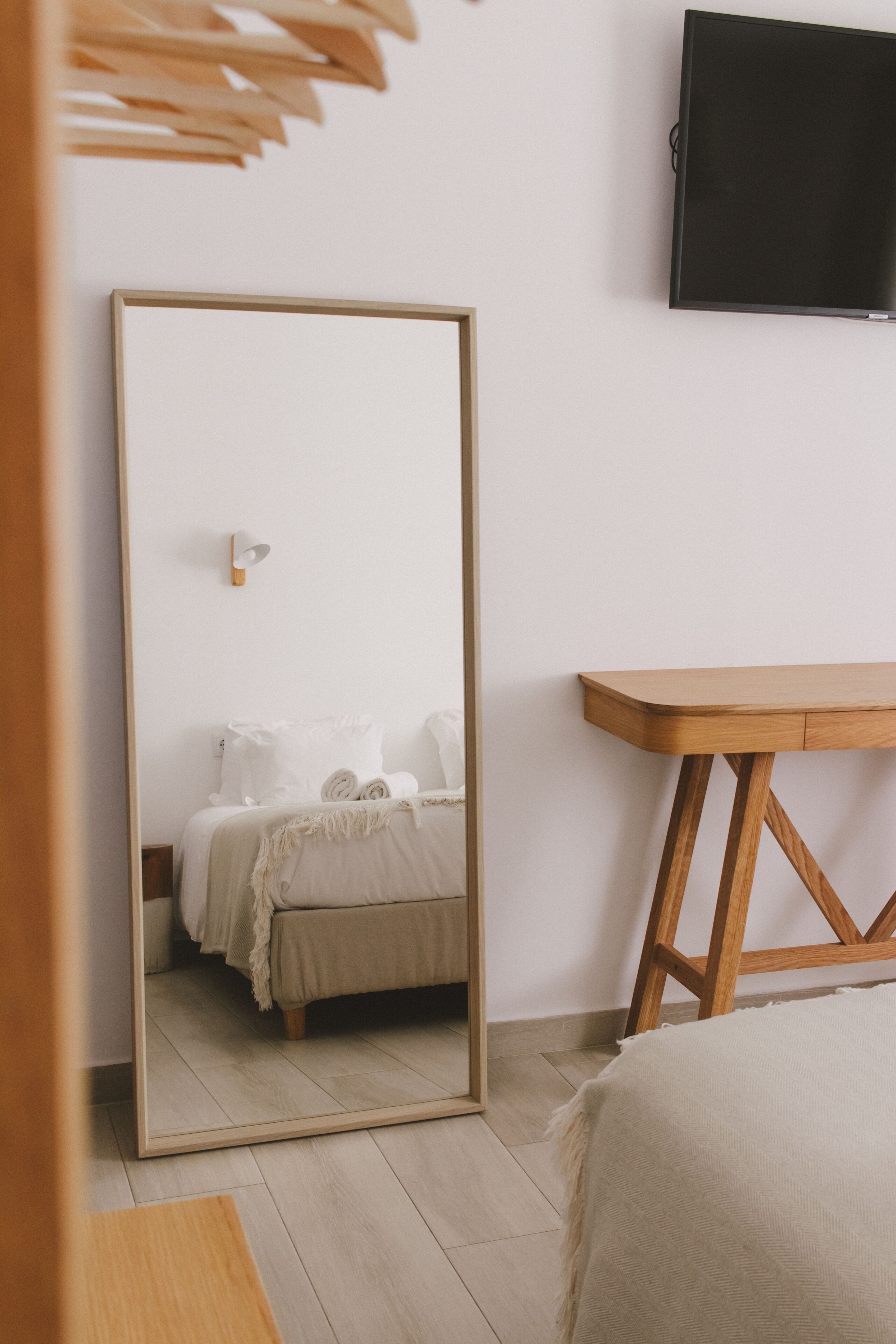Un dormitorio con un espejo grande, una cama con ropa de cama blanca, una mesita de noche de madera y un televisor de pantalla plana montado en la pared.