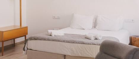 Quarto com cama, poltrona cinza, escrivaninha de madeira e mesa de cabeceira.