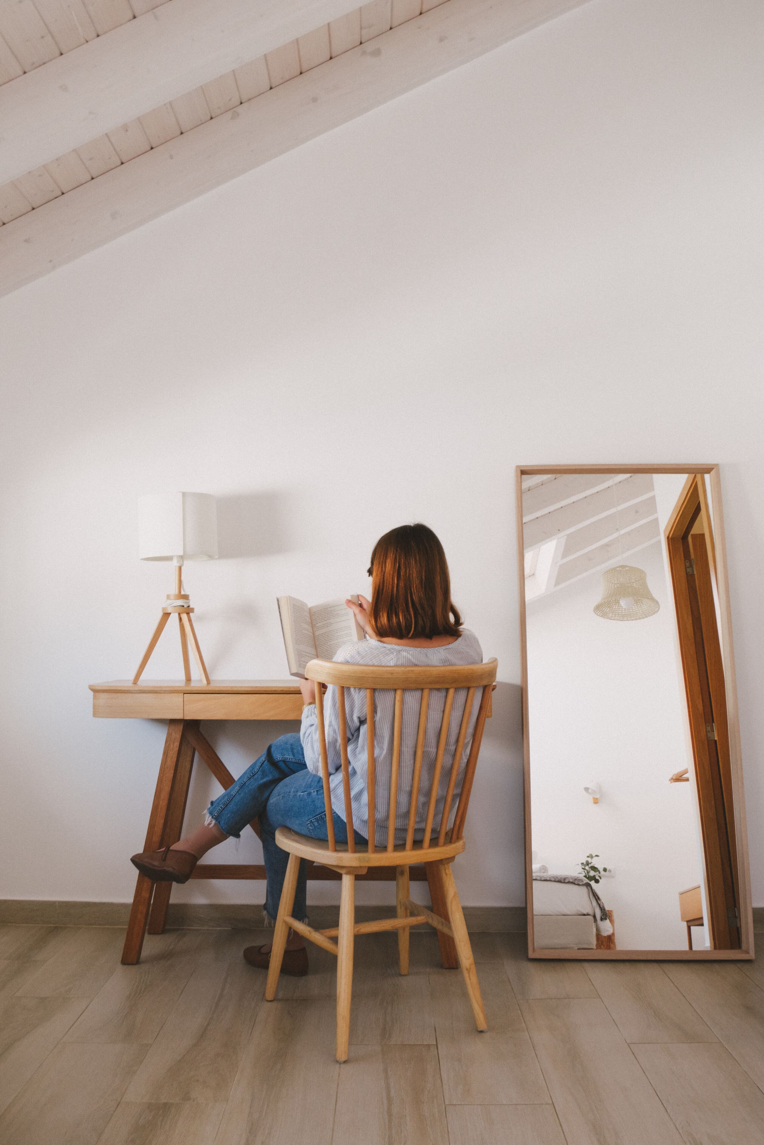 Una persona sentada en un escritorio de madera, con una lámpara, leyendo un libro, en una habitación con un espejo grande y piso de madera.