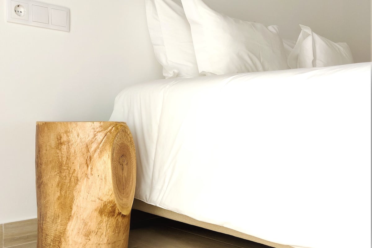 A bedroom with a bed, white bedding, a wooden nightstand, and a wall-mounted light fixture.