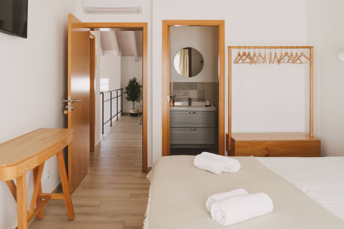 A hotel room with a wooden bed, a bedside table, a mirror, a wardrobe, and a bathroom visible through an open door.