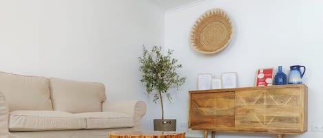 A living room with a beige sofa, a wooden coffee table, a potted plant, a woven wall hanging, and a wooden cabinet with decorative items.