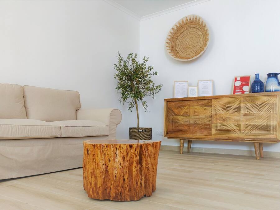 En stue med en beige sofa, et træ sofabord, en potteplante, et vævet vægophæng og et træskab med dekorative genstande.