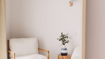 Una habitación minimalista con una silla de madera, una pequeña mesita de madera con un florero y una cama blanca.