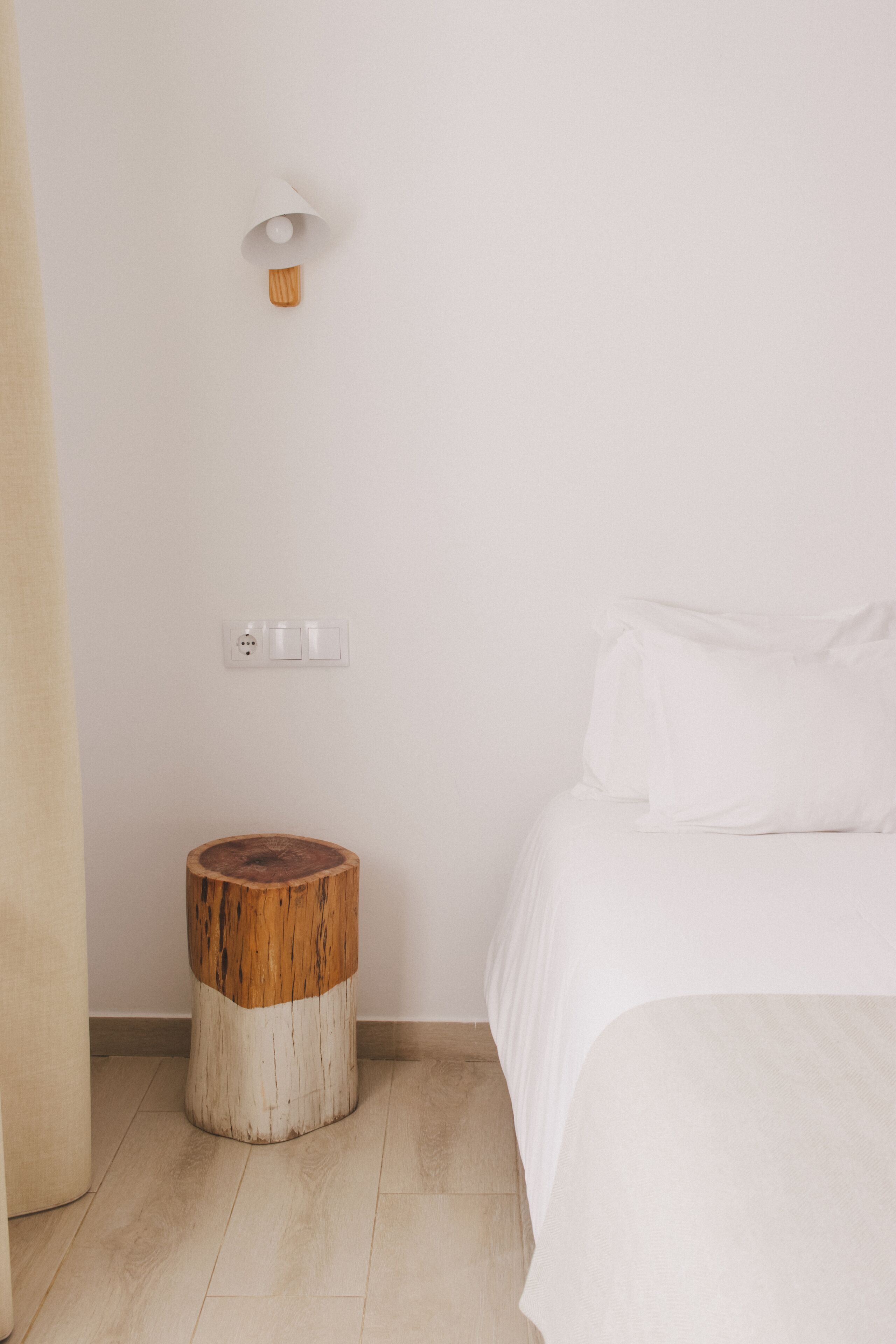 A minimalist bedroom with a wooden stool, a bedside table, and a wall-mounted light fixture.