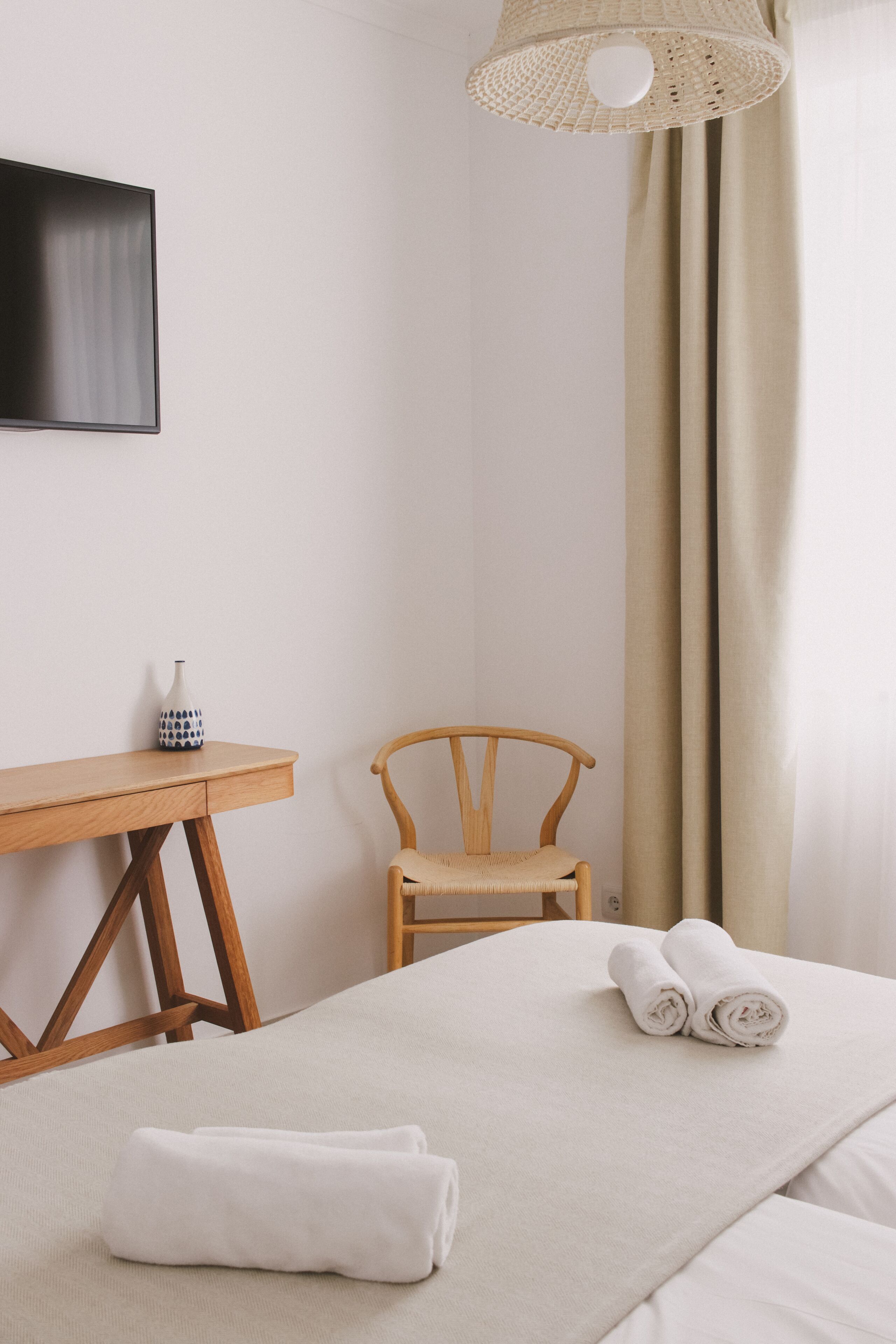 Ein ordentlich eingerichtetes Hotelzimmer mit einem Bett, einem Holzstuhl, einem kleinen Tisch mit einer Vase und einem Fernseher an der Wand.