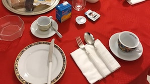 Aamiaiselle katettu pöytä, jossa on punainen pöytäliina, lautasia, aterimia, kulho täynnä hedelmiä, lasi maitoa ja erilaisia aamiaistarvikkeita.