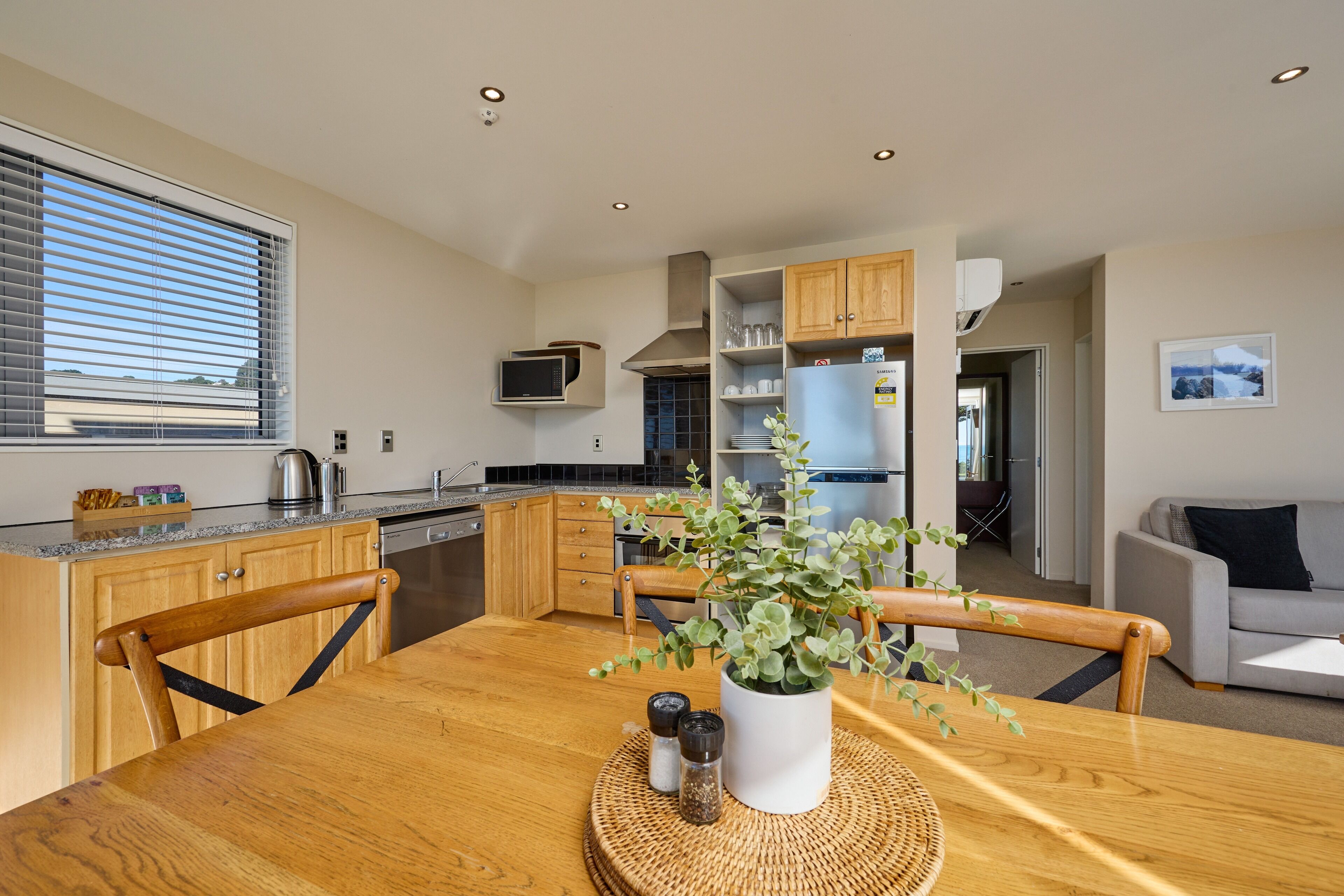 A modern kitchen with wooden cabinets, a dining table with a potted plant, and a living area with a sofa.
