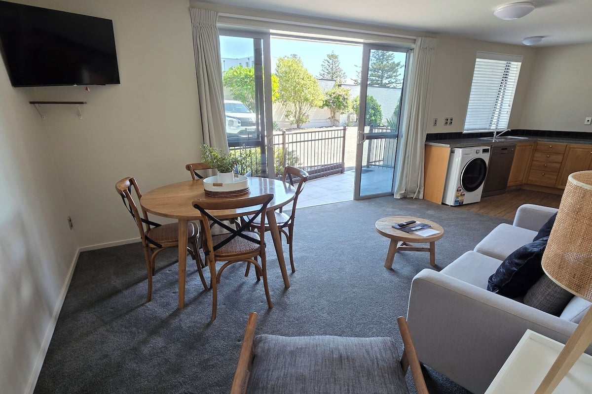 A modern living room with a dining area, a sofa, a TV mounted on the wall, and a sliding glass door leading to a balcony.