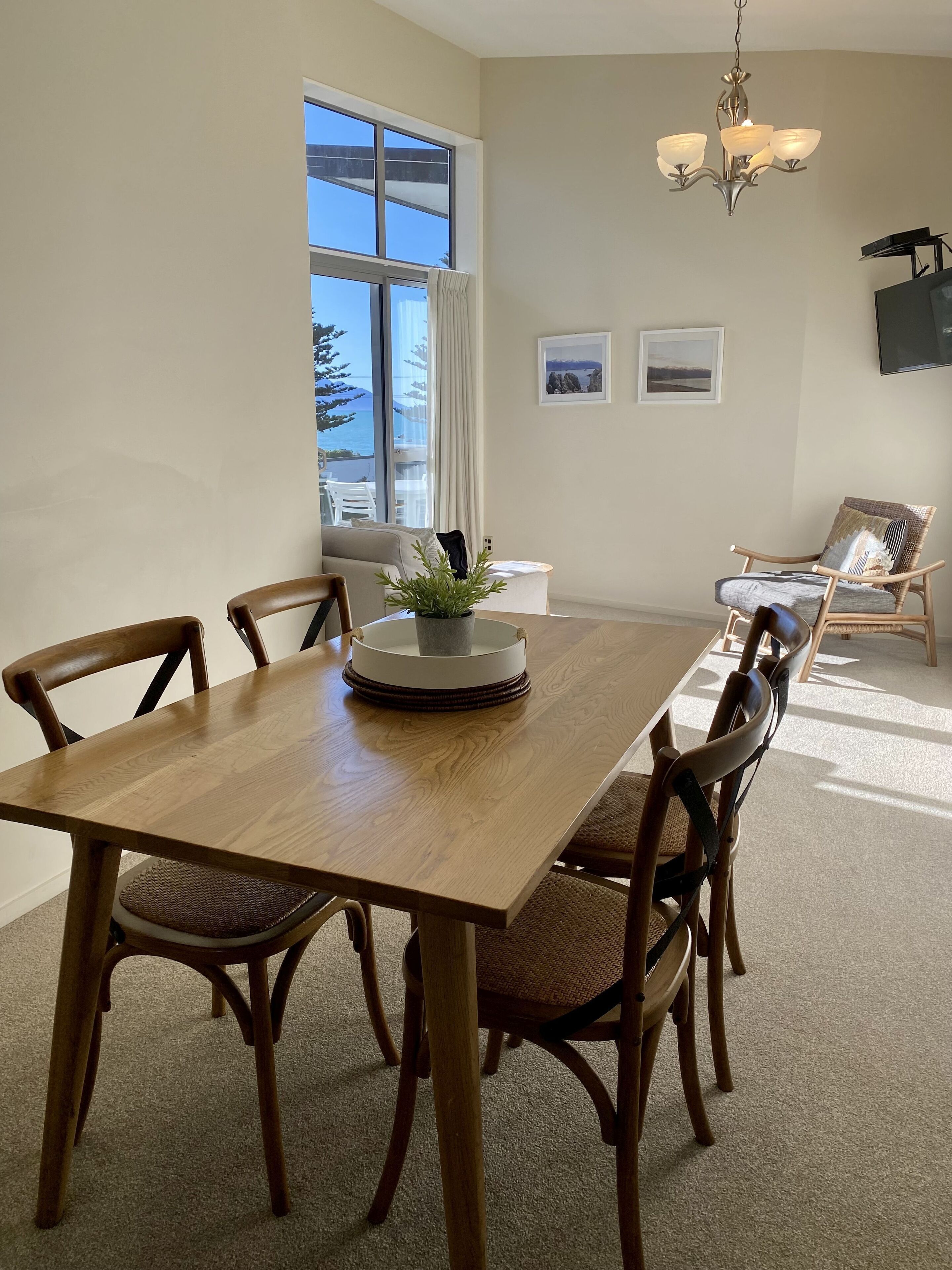 Un comedor con una mesa de madera, cuatro sillas, una planta en maceta y vistas del exterior a través de un gran ventanal.