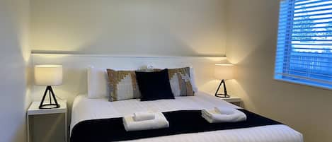 A hotel room with a large bed, two bedside tables with lamps, a window with blinds, and a view of trees outside.