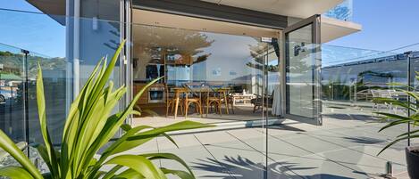 A modern outdoor dining area with glass walls, a table, chairs, and potted plants.