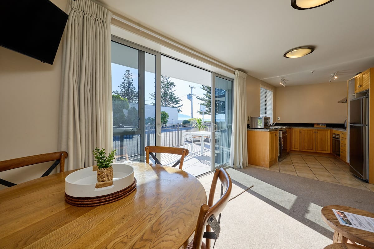 A modern kitchen with a large island, a dining area with a wooden table, and a sliding glass door leading to a balcony with a view of trees and a building.