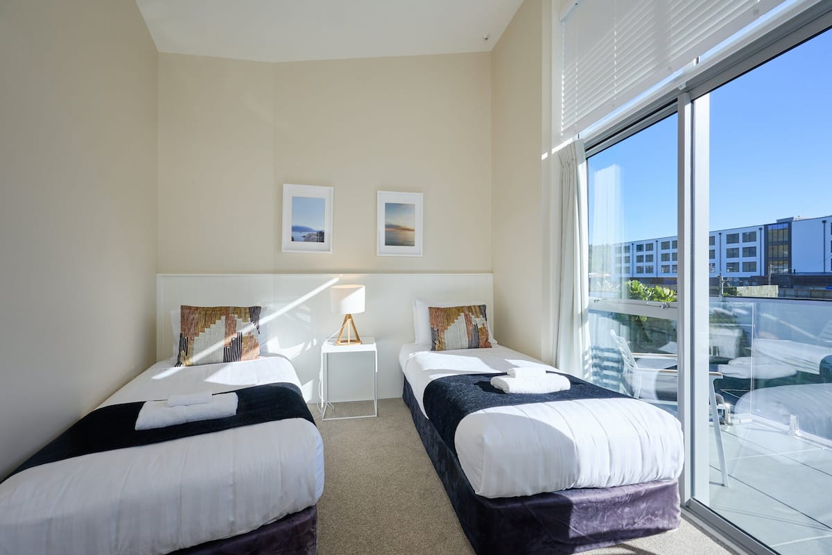 A hotel room with two beds, a small bedside table with a lamp, and a large window offering a view of the city.