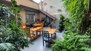 Amplio espacio interior con techo alto, mesas y sillas de madera, una escalera y abundante vegetación.