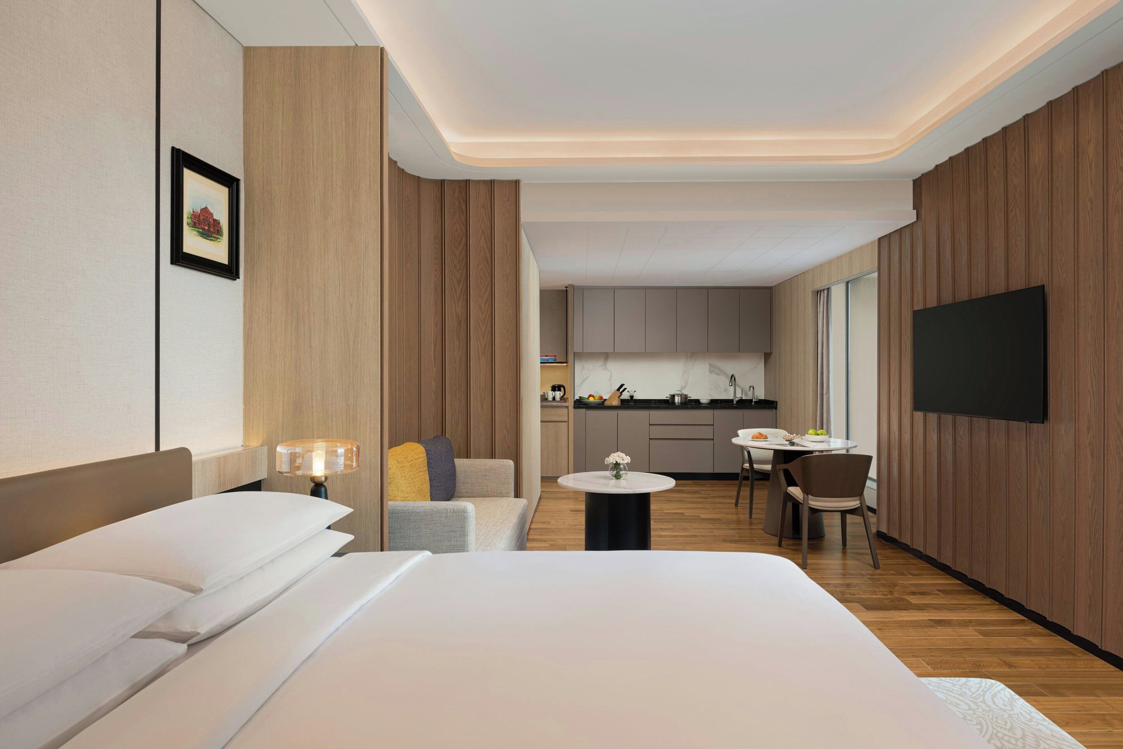 Une chambre d’hôtel avec un lit, une petite table, une chaise, un téléviseur et, au fond, une kitchenette.