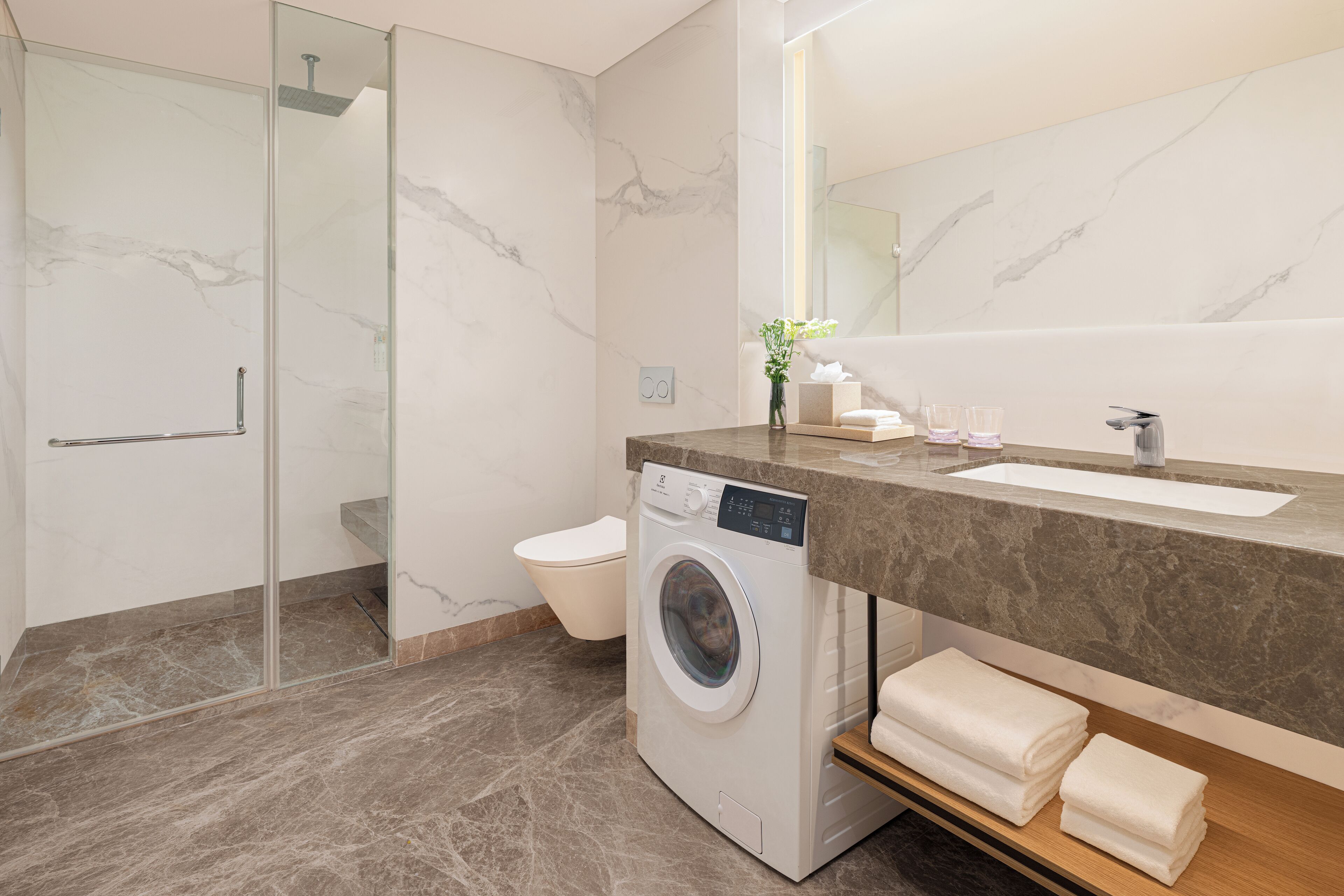 A modern bathroom with a marble floor, a washing machine, a sink, and a glass shower enclosure.